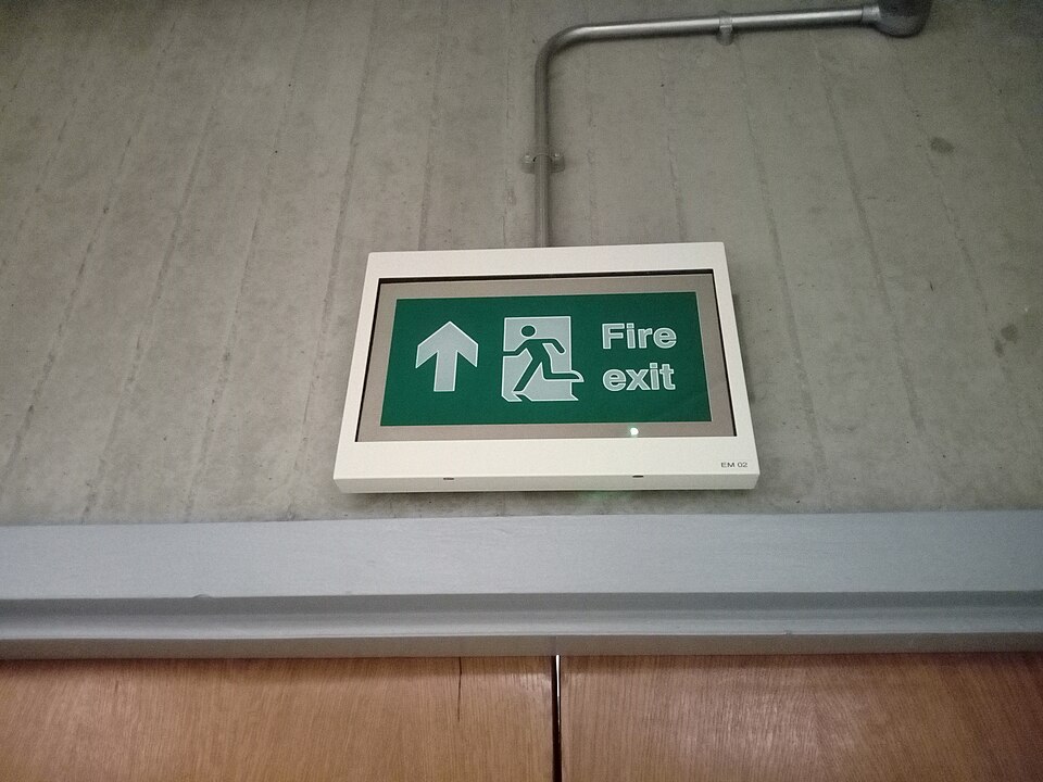 A maintained fire exit sign above a doorway in a UK building, lit and showing the direction of escape